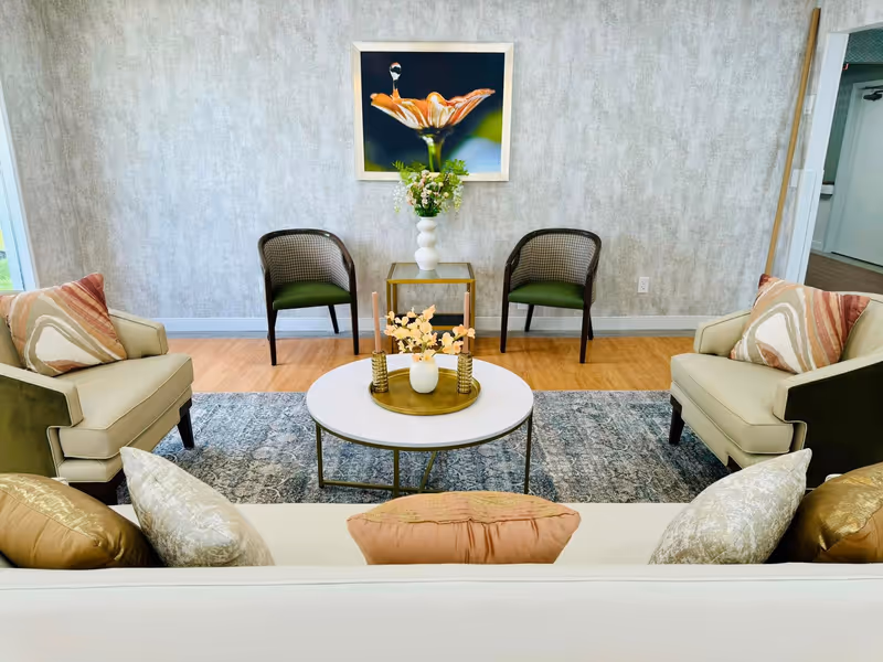 A cozy living room area with a white sofa adorned with multiple pillows in gold, cream, and peach tones. In front of the sofa is a round white coffee table with a gold tray holding a small vase with flowers and two gold candle holders. Two beige armchairs with patterned pillows face the sofa. Behind the coffee table, there are two dark wood chairs with green cushions and a small glass side table with a white vase of flowers. A framed close-up photograph of an orange flower with a water droplet hangs on the textured gray wall. The floor is a combination of wood and a patterned area rug.