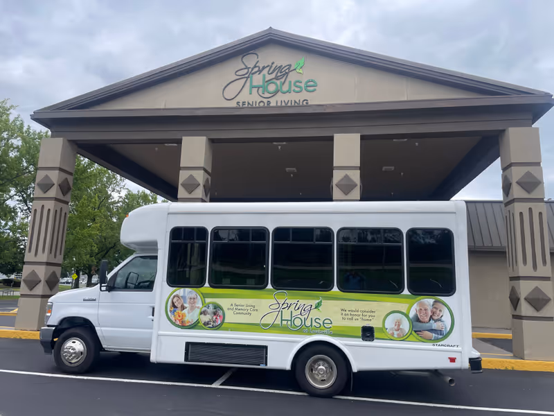 A white shuttle bus parked under the entrance canopy of The Spring House at Louisville senior living facility. The bus has a green and white advertisement on its side featuring smiling elderly people and text about the senior living and memory care community. The building entrance has a sign that reads 'Spring House Senior Living'.