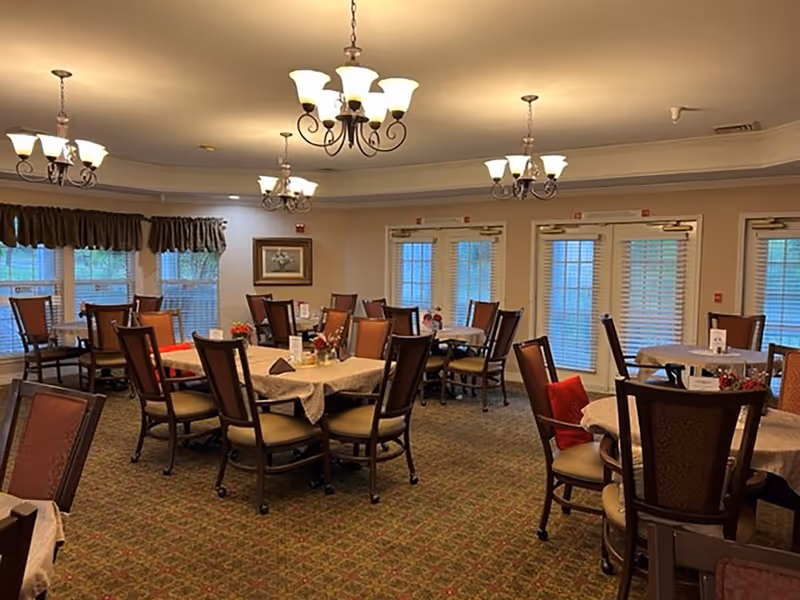 A dining room with multiple round tables covered with beige tablecloths, each surrounded by wooden chairs with cushioned seats and backs. The room is well-lit with several ornate chandeliers hanging from the ceiling. Windows with brown valances and glass doors with blinds line the walls, and there are small floral centerpieces on the tables.