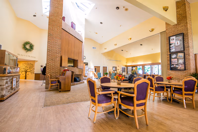 A spacious and well-lit common area in a senior living facility with multiple round tables surrounded by wooden chairs with purple cushions. The room features a high ceiling with skylights, a large brick fireplace, a piano, and various decorations including framed photos, plants, and wall art. The flooring is a combination of wood and carpet, and large windows allow natural light to fill the space.
