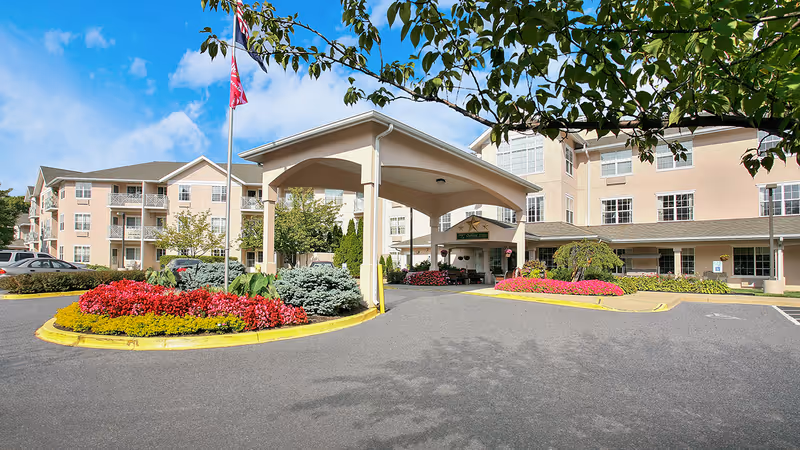 Front entrance of a multi-story senior living building with a covered porte-cochere, circular landscaped flowerbeds, and parked cars.
