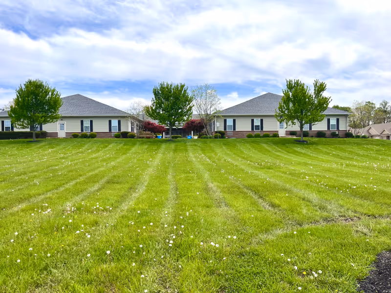 Wide view of a single-story senior living facility building with beige siding and brick accents, surrounded by a well-maintained green lawn with scattered white flowers and several trees under a partly cloudy sky.