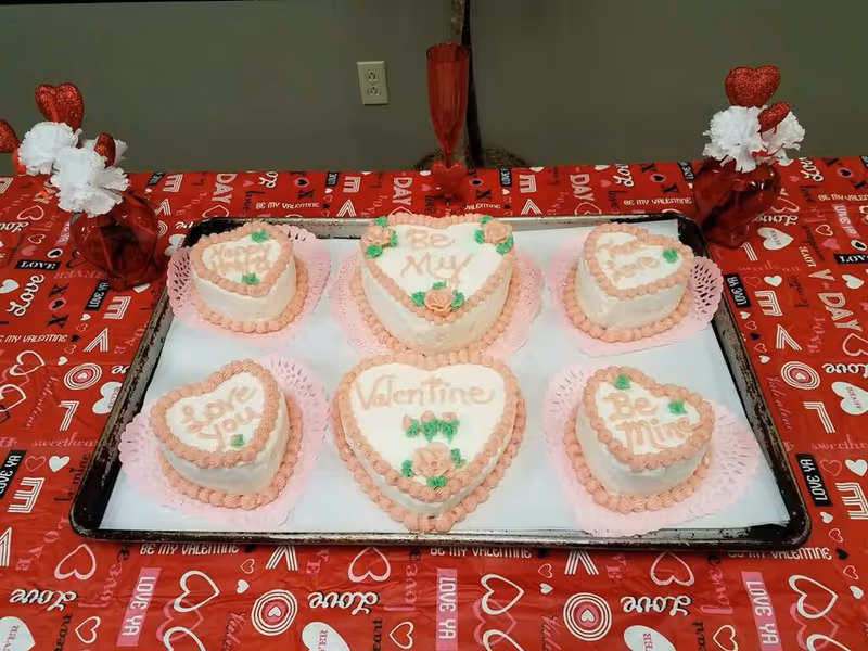 A tray of six heart-shaped Valentine's cakes with pink frosting arranged on a red Valentine-themed tablecloth with small heart centerpieces.