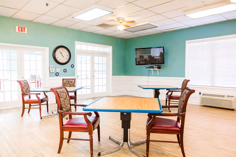 A bright room with light wood flooring and teal walls featuring three tables with wooden chairs that have red cushioned seats and floral backs. A wall clock and a mounted flat-screen TV are visible, along with large windows with blinds and a glass door with an exit sign above it.