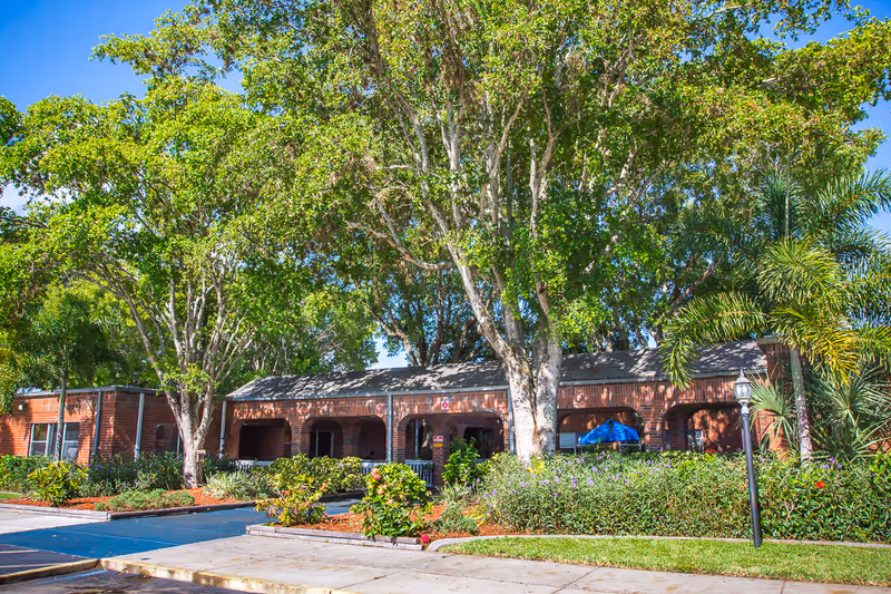 Front exterior of a single-story brick rehab and wellness center with large shade trees, landscaped beds, and a driveway.
