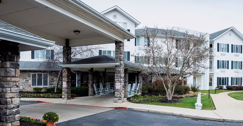 Front entrance of a multi-story senior living building with a covered porte-cochere, stone columns, rocking chairs, and landscaped grounds.