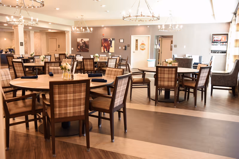 A spacious dining room with multiple round and rectangular tables surrounded by wooden chairs with plaid upholstery. The tables are set with small flower arrangements and dark napkins. The room features modern chandeliers, light-colored walls, and wood flooring with a patterned design. In the background, there is a beverage dispenser and some seating areas near windows with curtains.