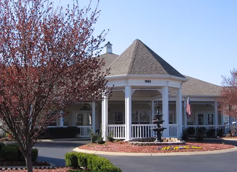 Front entrance of a single-story senior living building with a white gazebo-style portico marked "981", a fountain in a circular driveway, landscaping and an American flag.