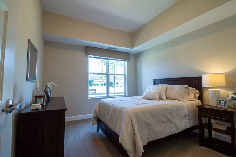 Bright, furnished bedroom with a bed, nightstand and lamp, dresser, and a sunlit window.