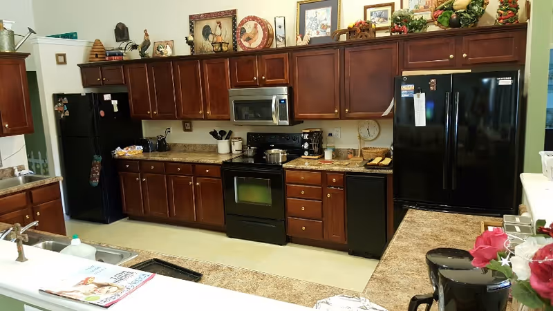 A kitchen with dark wooden cabinets, black appliances including a refrigerator, stove, microwave, and dishwasher. The countertops are granite with various kitchen items such as a coffee maker, utensils, and decorative pieces on top of the cabinets. There is a double sink in the foreground with a magazine placed nearby and a vase with red and white flowers on the right side.