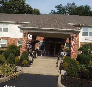 Entrance of Creekside Senior Apartments featuring a covered porch with brick pillars, an American flag, and neatly trimmed bushes along the walkway leading up to the double doors.