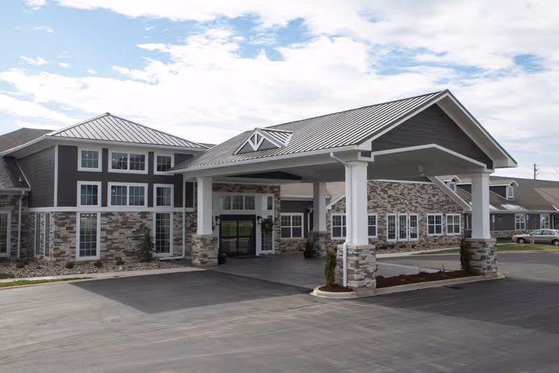 Exterior view of Walker's Trail Senior Living facility showing a large covered entrance with stone and siding facade, multiple windows, and a paved driveway under a partly cloudy sky.