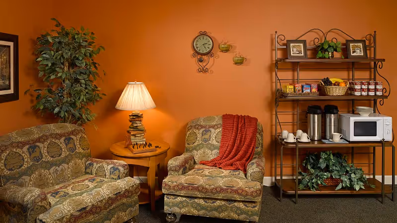 A cozy sitting area with two patterned armchairs, one draped with a red throw blanket, a wooden side table with a lamp and a chessboard, a tall leafy plant, and a metal shelving unit holding snacks, coffee dispensers, a microwave, cups, plates, and decorative plants against an orange wall with a clock and framed pictures.