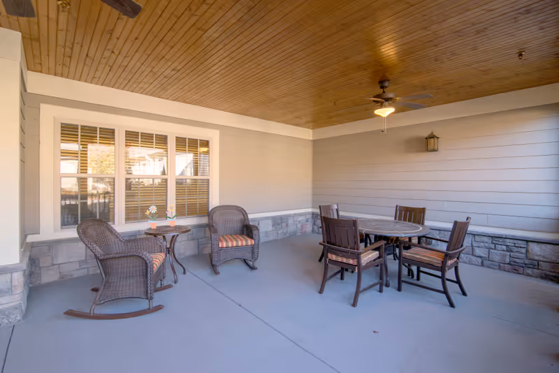 Covered patio with wicker rocking chairs and a round dining table with four chairs under a wood-paneled ceiling and ceiling fan.