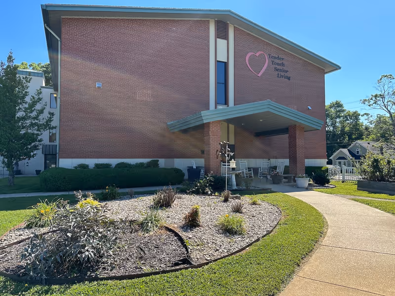 Brick senior living building front with a covered entrance, rocking chairs on the porch, landscaped circular planter, and a heart logo reading 'Tender Touch Senior Living'.