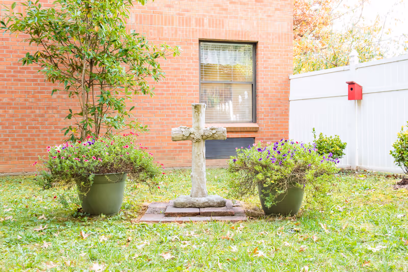 Outdoor garden area with a stone cross monument centered between two large green pots filled with flowering plants. Behind the cross is a brick building wall with a window, and to the right is a white fence with a red birdhouse attached.