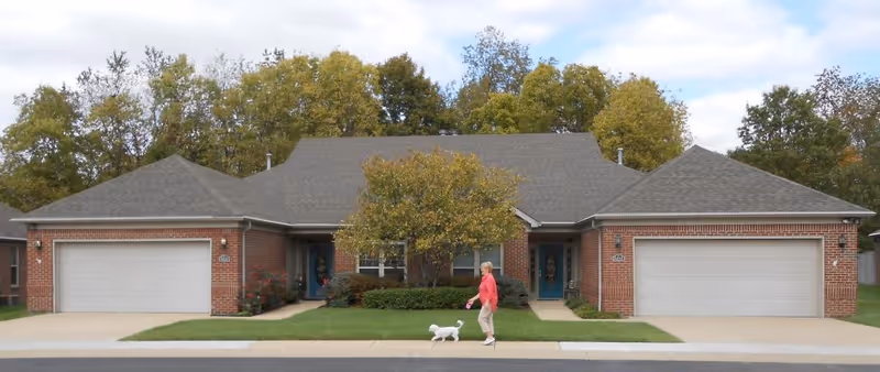 Brick duplex-style building with two garages and a central entrance, a tree and shrubs in front, and a person walking a small white dog on the sidewalk.