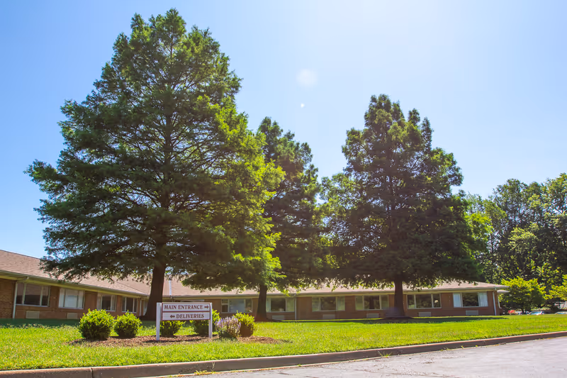 Exterior view of a single-story brick building with a sloped roof, surrounded by large green trees and a well-maintained lawn under a clear blue sky. A sign in front indicates directions to the main entrance and deliveries.