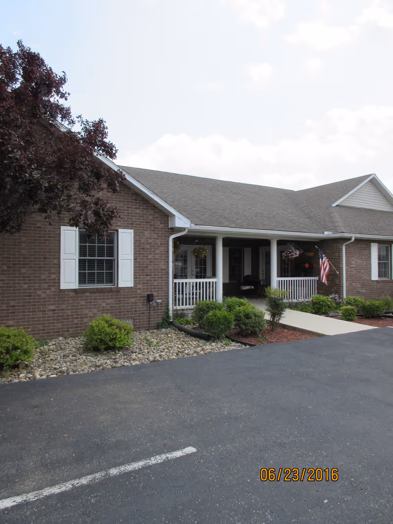 Exterior view of a single-story brick building with white window shutters and a covered porch with white railings. There are small bushes and landscaping rocks along the front, an American flag hanging near the porch, and a paved driveway in front. The sky is partly cloudy.