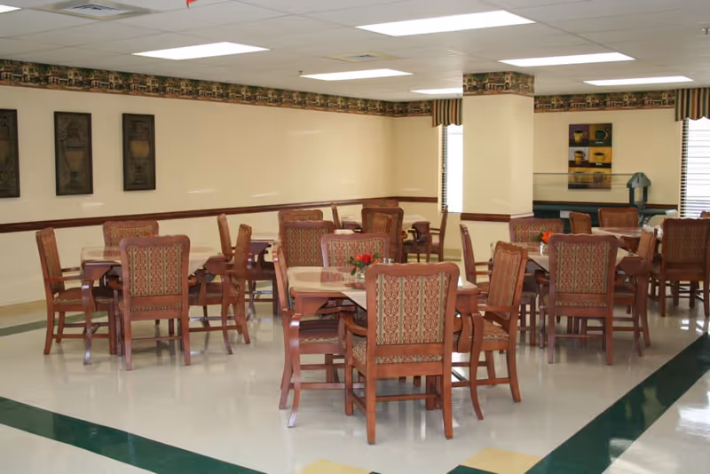 Dining room with several wooden tables and upholstered chairs arranged on a tiled floor under a drop ceiling, each table topped with a small floral centerpiece.