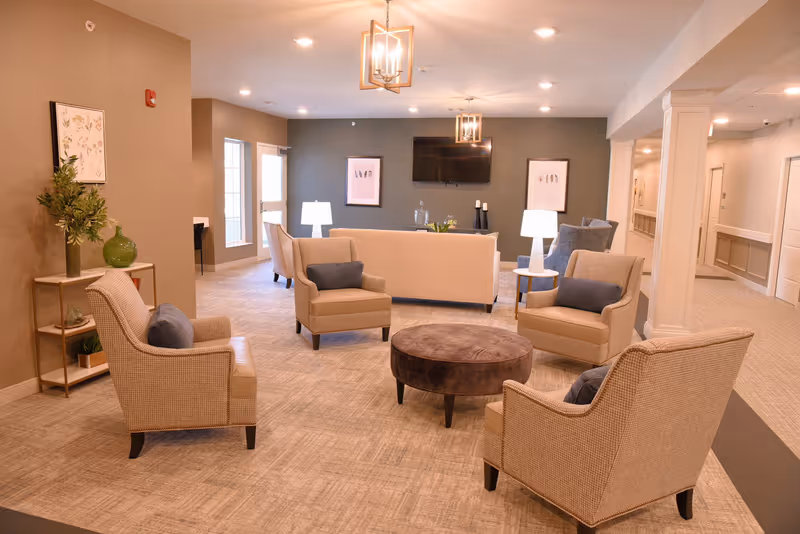 A cozy senior living facility common area with beige armchairs and a sofa arranged around a round brown ottoman. The room features soft lighting from ceiling fixtures and table lamps, neutral-colored walls with framed artwork, and a flat-screen TV mounted on a dark accent wall. There is a small table with decorative plants on the left and a hallway visible on the right.