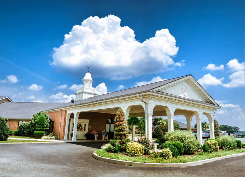 Exterior view of The Grandview Nursing and Rehabilitation Facility showing a covered entrance with white columns, well-maintained landscaping with bushes and small trees, and a bright blue sky with large white clouds.