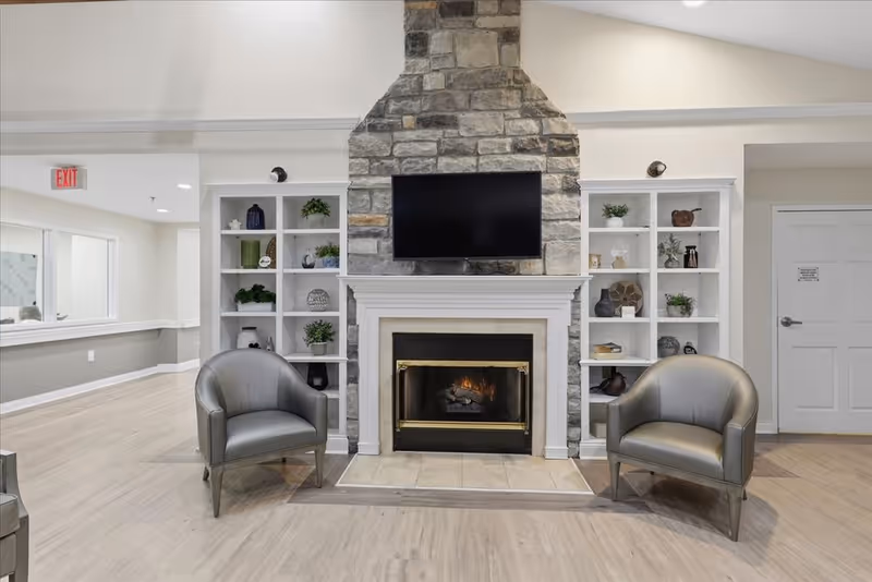 Bright interior living room with stone fireplace, TV, built-in shelves, and two gray chairs.