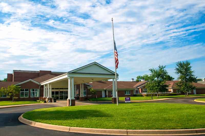 Exterior view of the Episcopal Church Home building with a covered entrance, a flagpole with an American flag at half-mast, green lawn, trees, and a partly cloudy blue sky.