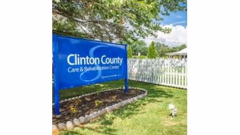 Outdoor view of a blue sign for Clinton County Care & Rehabilitation Center placed in a landscaped area with grass, flowers, and a white picket fence in the background under a partly cloudy sky.