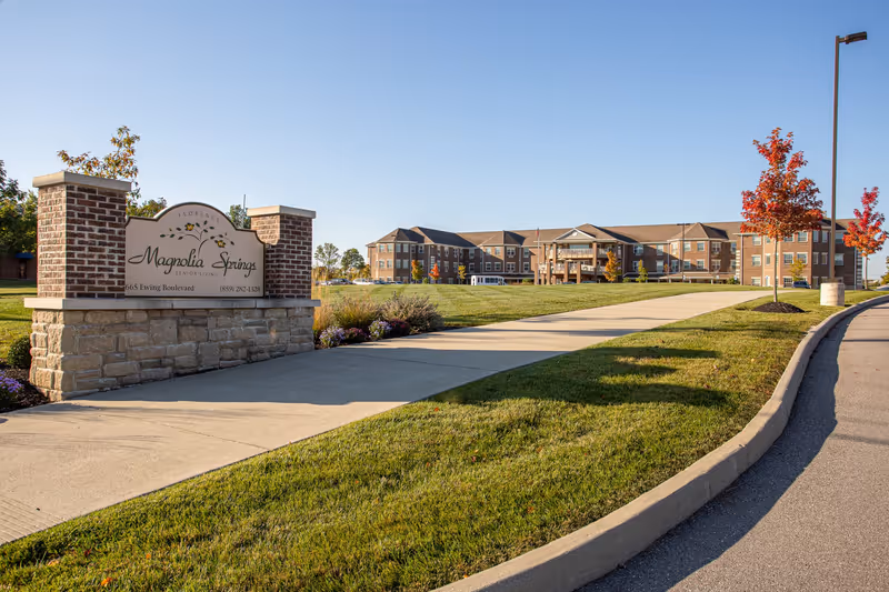Wide exterior view of Magnolia Springs Florence senior living facility with a large brick and stone sign in the foreground on the left, a paved walkway leading to the building, green lawns, and trees with autumn foliage under a clear blue sky.