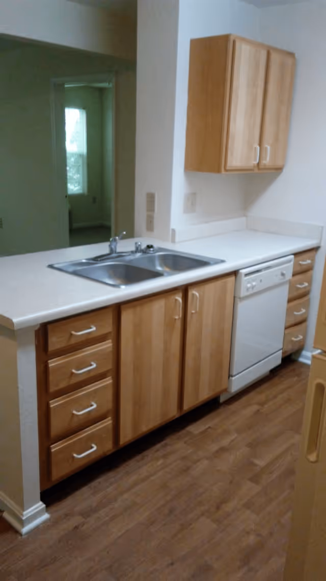A kitchen area with wooden cabinets and drawers, a double stainless steel sink, a white dishwasher, and a white countertop. The floor is wooden, and there is an open doorway leading to another room with a window.