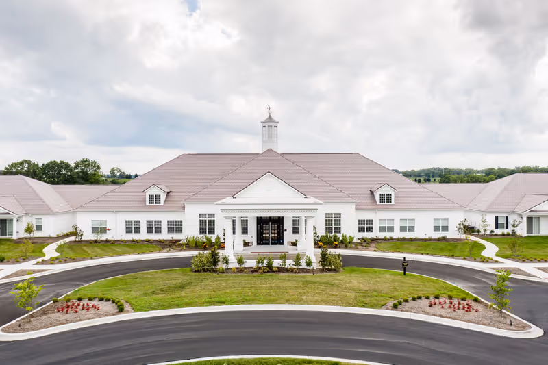 Front exterior view of a large, single-story building with a white facade and a brown roof, featuring a central entrance with columns and a cupola on top. The building is surrounded by landscaped grass, small plants, and a circular driveway.