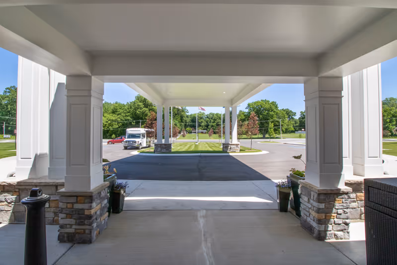 View from under a covered entrance area of a building looking out onto a driveway with a parked vehicle, green lawn, trees, and an American flag in the distance on a sunny day.