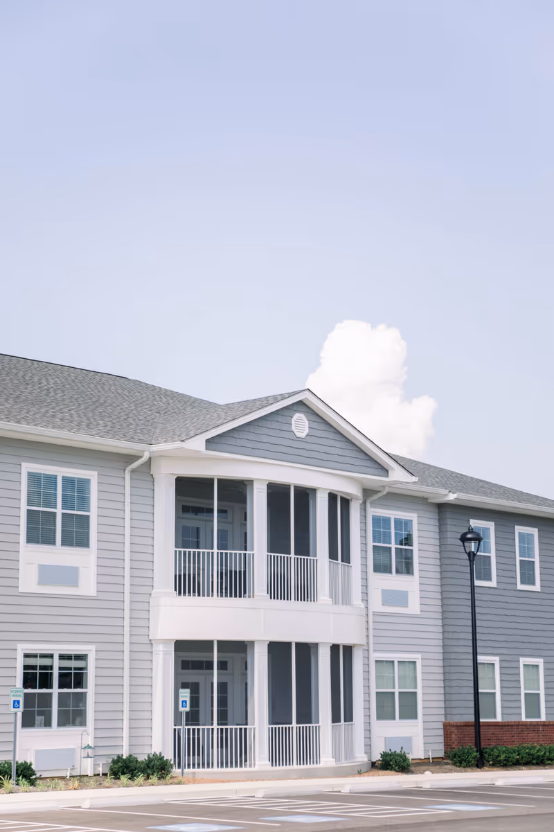 Exterior view of a two-story senior living facility building with gray siding, white trim, and a covered balcony. The building has multiple windows and a parking area with handicap parking spaces in front. A street lamp is visible on the right side.