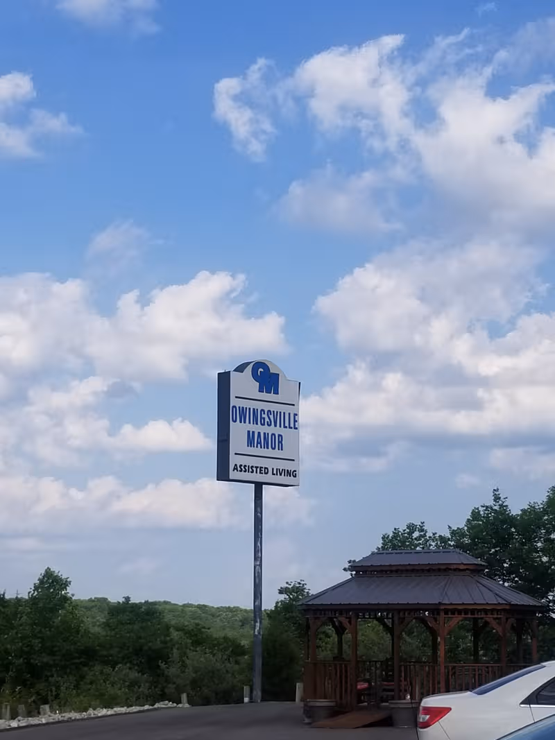 Outdoor scene featuring Owingsville Manor sign and gazebo against blue sky.