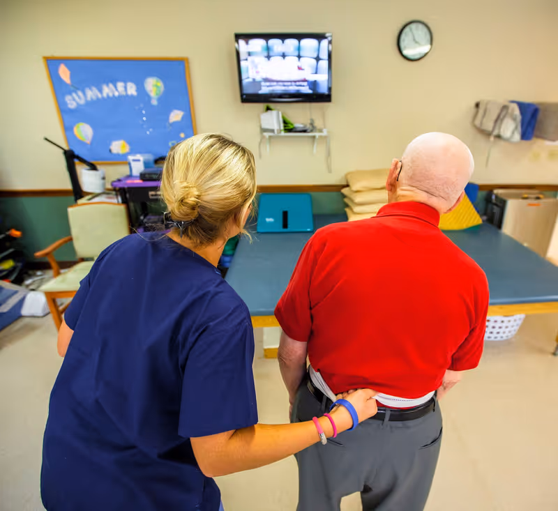 A caregiver in navy scrubs is assisting an elderly man in a red shirt by measuring his waist with a tape measure in a room with exercise equipment and a bulletin board labeled 'SUMMER'.
