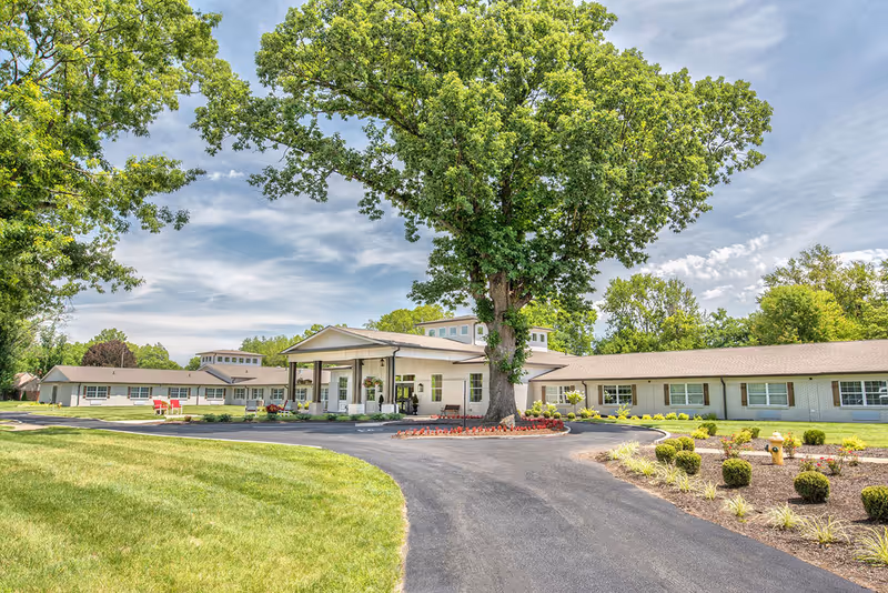 Front exterior of a single-story senior living facility with a covered entrance, large tree, circular driveway and landscaped lawn.
