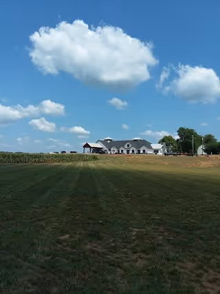 A large white building with a dark roof situated in the distance on a grassy field under a blue sky with scattered clouds.
