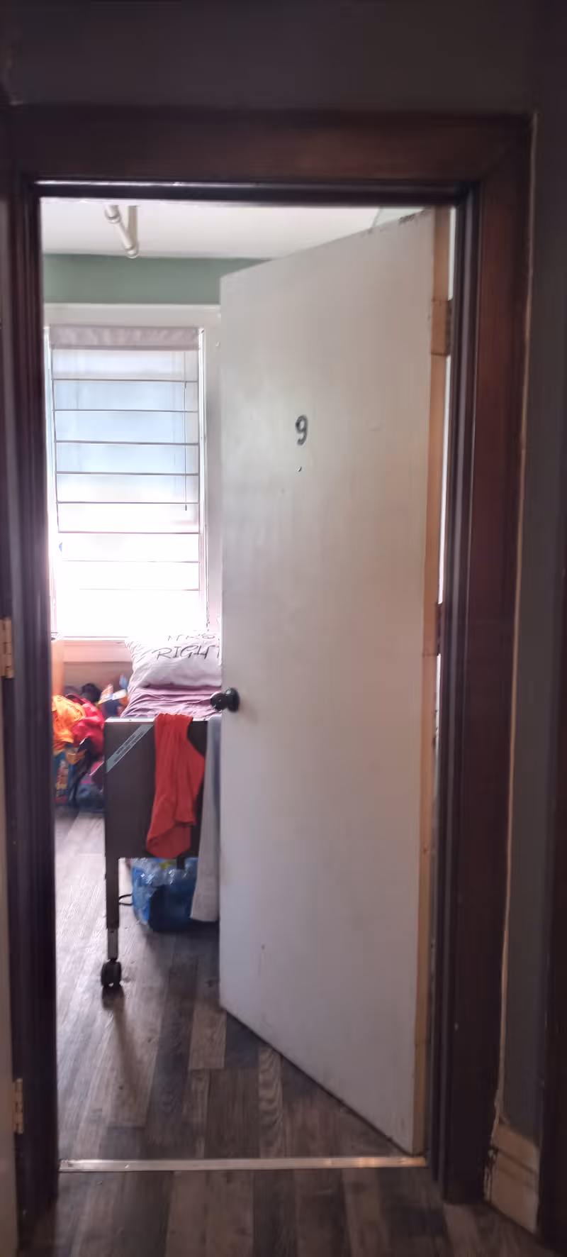 Open door reveals a bedroom with a bed, clothes, and window blinds.
