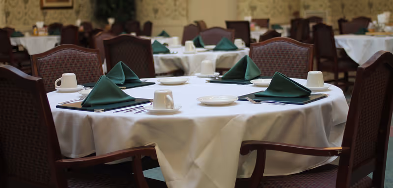 A dining room with round tables covered in white tablecloths, each set with green folded napkins, white cups on saucers, plates, and silverware. The chairs around the tables have wooden frames and patterned upholstery.