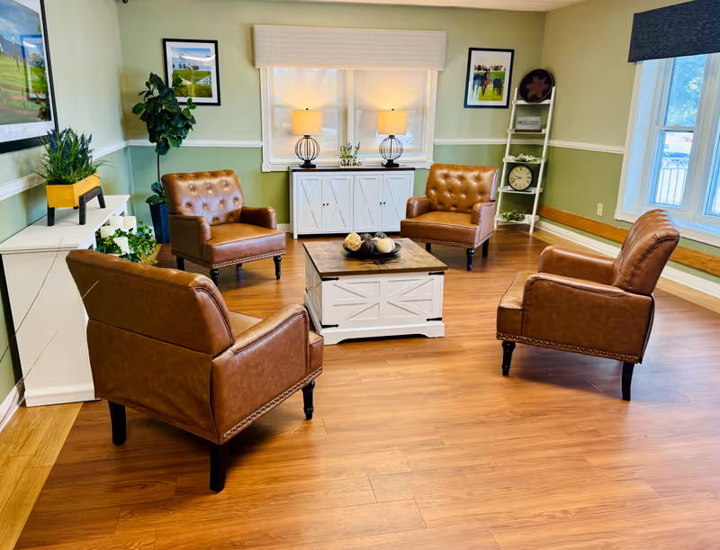 A cozy sitting area with four brown leather armchairs arranged around a white wooden coffee table with decorative items. The room has light green walls with white trim, wooden flooring, two windows with white blinds, a white cabinet with two lamps on top, framed pictures on the walls, and a white ladder shelf with decorative items including a clock and a welcome sign.