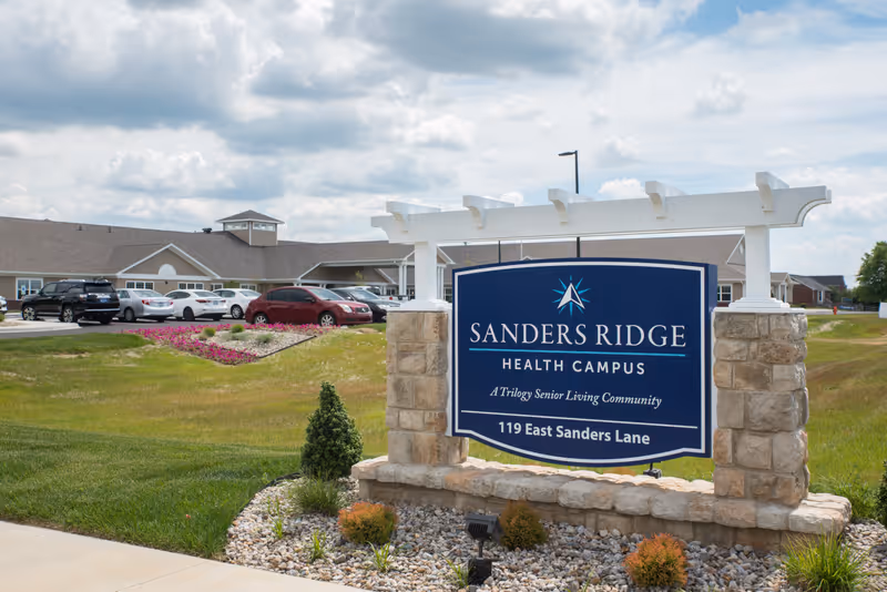Exterior view of Sanders Ridge Health Campus sign with a landscaped area and parked cars in front of a large building under a partly cloudy sky.