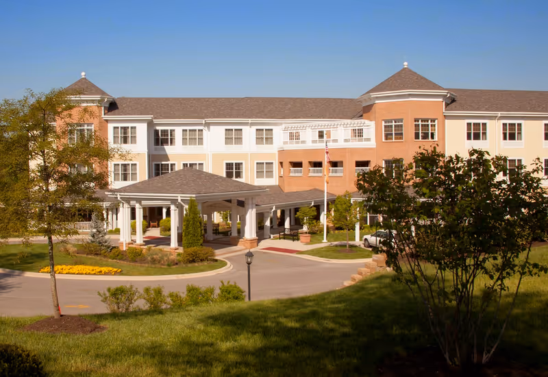 Exterior view of a large senior living facility building with a covered entrance, multiple windows, and two turret-like structures on each end. The building is surrounded by landscaped greenery, trees, and a circular driveway under a clear blue sky.