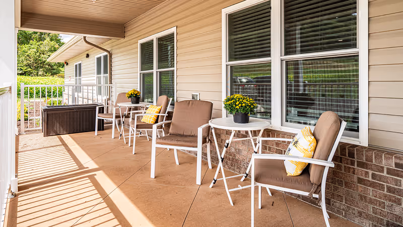 A covered outdoor patio area with beige siding and brick wall, featuring white metal chairs with brown cushions and yellow patterned pillows, small white round tables with potted yellow flowers, and a storage bench. The patio is well-lit with natural sunlight and overlooks a green garden area.