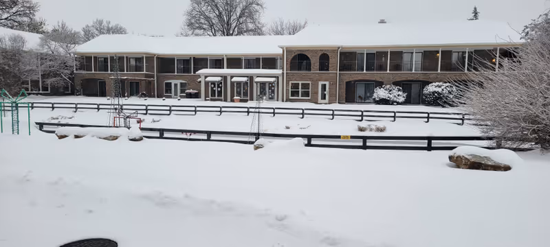 Snow-covered courtyard and fenced garden area in front of a two-story brick assisted living building with balconies and walkways.