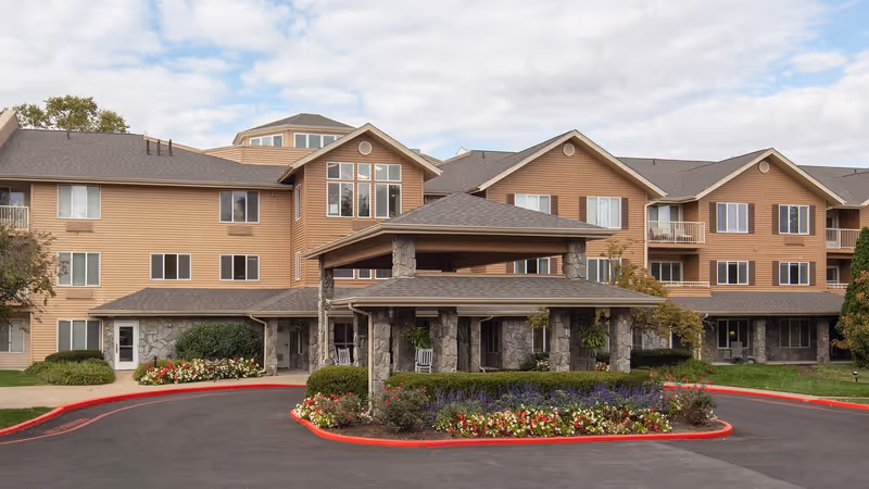 Exterior view of Holiday Hartland Hills senior living facility showing a multi-story building with beige siding and stone accents. The entrance features a covered drop-off area supported by stone pillars, surrounded by landscaped flower beds and greenery. The sky is partly cloudy.