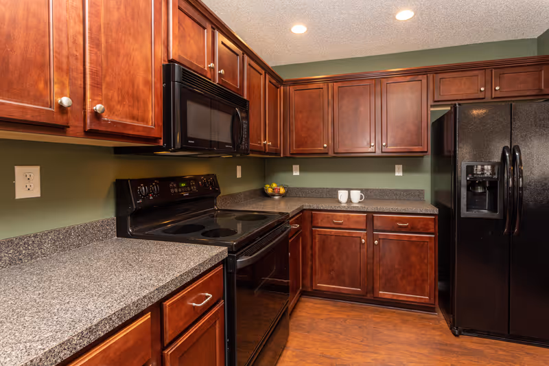 A kitchen with dark wood cabinets, black appliances including a stove, microwave, and refrigerator, and gray speckled countertops. The walls are painted green and the floor is wood. Two white mugs and a bowl of fruit are on the countertop.