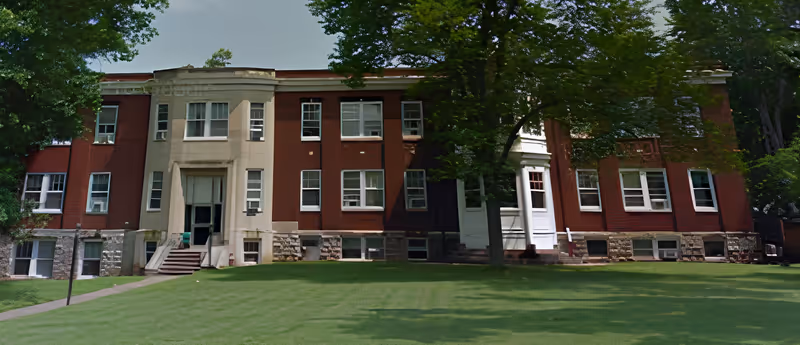 Front view of a two-story red brick institutional building with a white central entrance, many windows, and a large grassy lawn with trees.