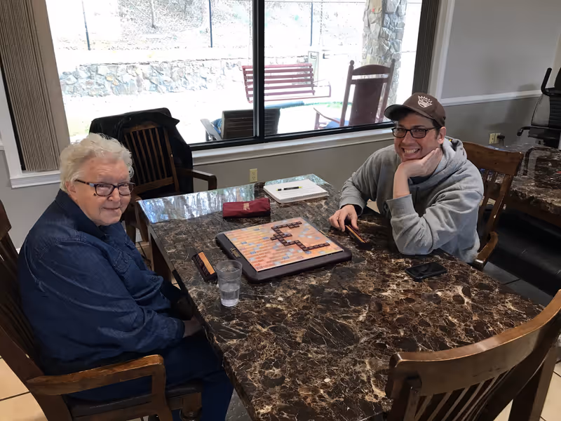 Two people sitting at a marble-top table playing a word board game near a large window.