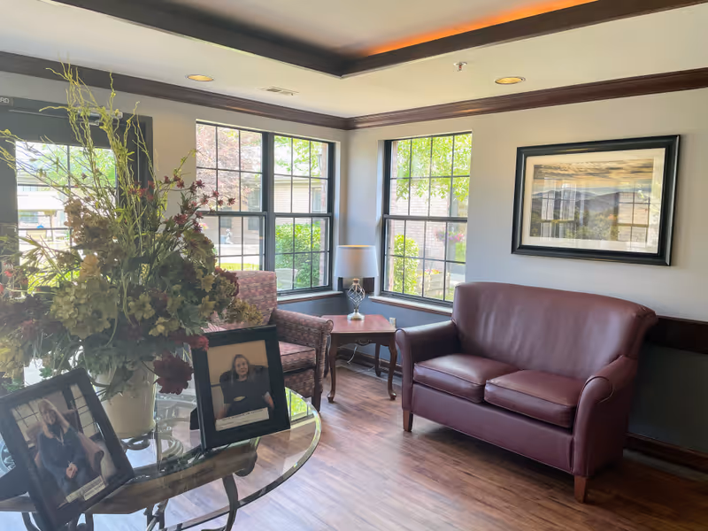 A cozy sitting area in a nursing and rehabilitation facility with a maroon leather loveseat, a patterned armchair, a wooden side table with a lamp, and large windows letting in natural light. A glass table in the foreground holds a large floral arrangement and two framed photographs of staff members. A framed landscape picture hangs on the wall.